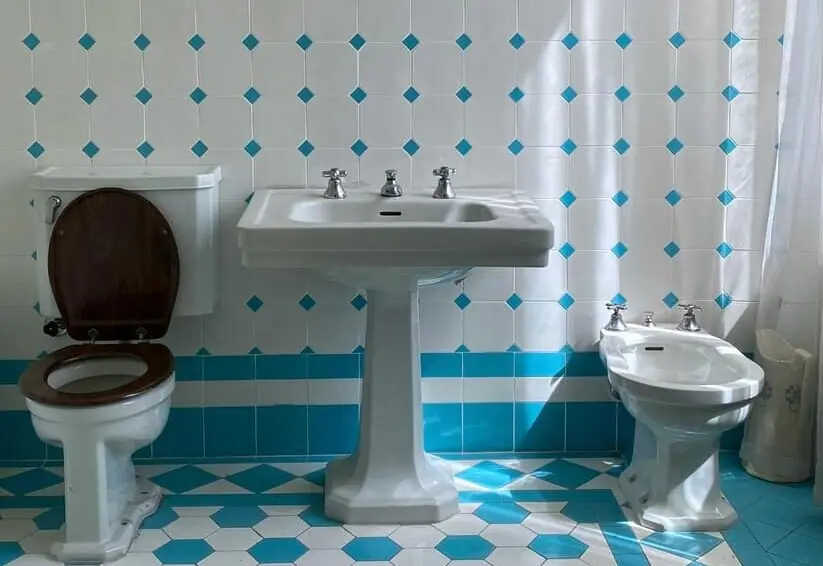 Klassieke badkamer met een staand toilet met houten zitting, een wastafel op zuil en een bidet tegen een wit-blauw betegelde wand, ter illustratie van kit- & voegonderhoud voor badkamers om vochtschade te voorkomen.