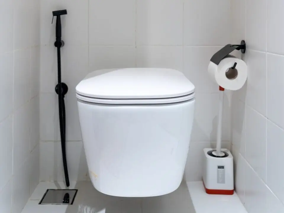 Modern hangend toilet met een zwart bedieningspaneel en handdouche in een wit betegelde toiletruimte, representatief voor een wandclosetmontage waarbij een sanibroyeur voor onderhoud achter de wand is geplaatst.
