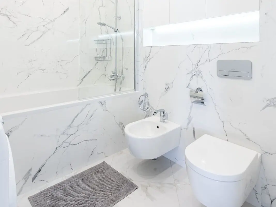 Modern hangend toilet en bidet tegen een wit marmeren wand in een strakke badkamer met ligbad, ter illustratie van een wandcloset waarin een sanibroyeur voor onderhoud is ingebouwd.